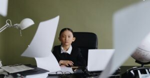 A child in formal wear sits at a desk surrounded by flying papers, depicting creativity and ambition.